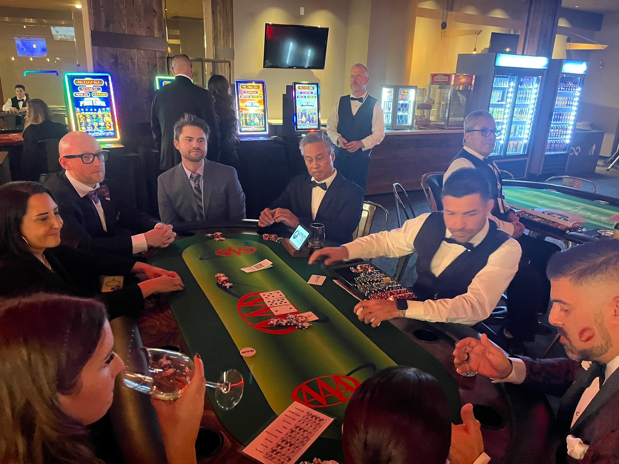 Guests playing poker at custom AAA logo branded casino table felt with slot machines in background