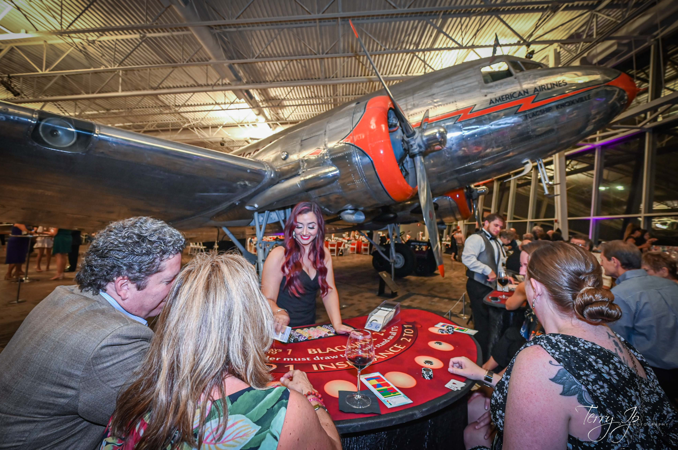 Dealer and guests at a blackjack table beneath a vintage American Airlines plane at a Dallas Casino Party USA event