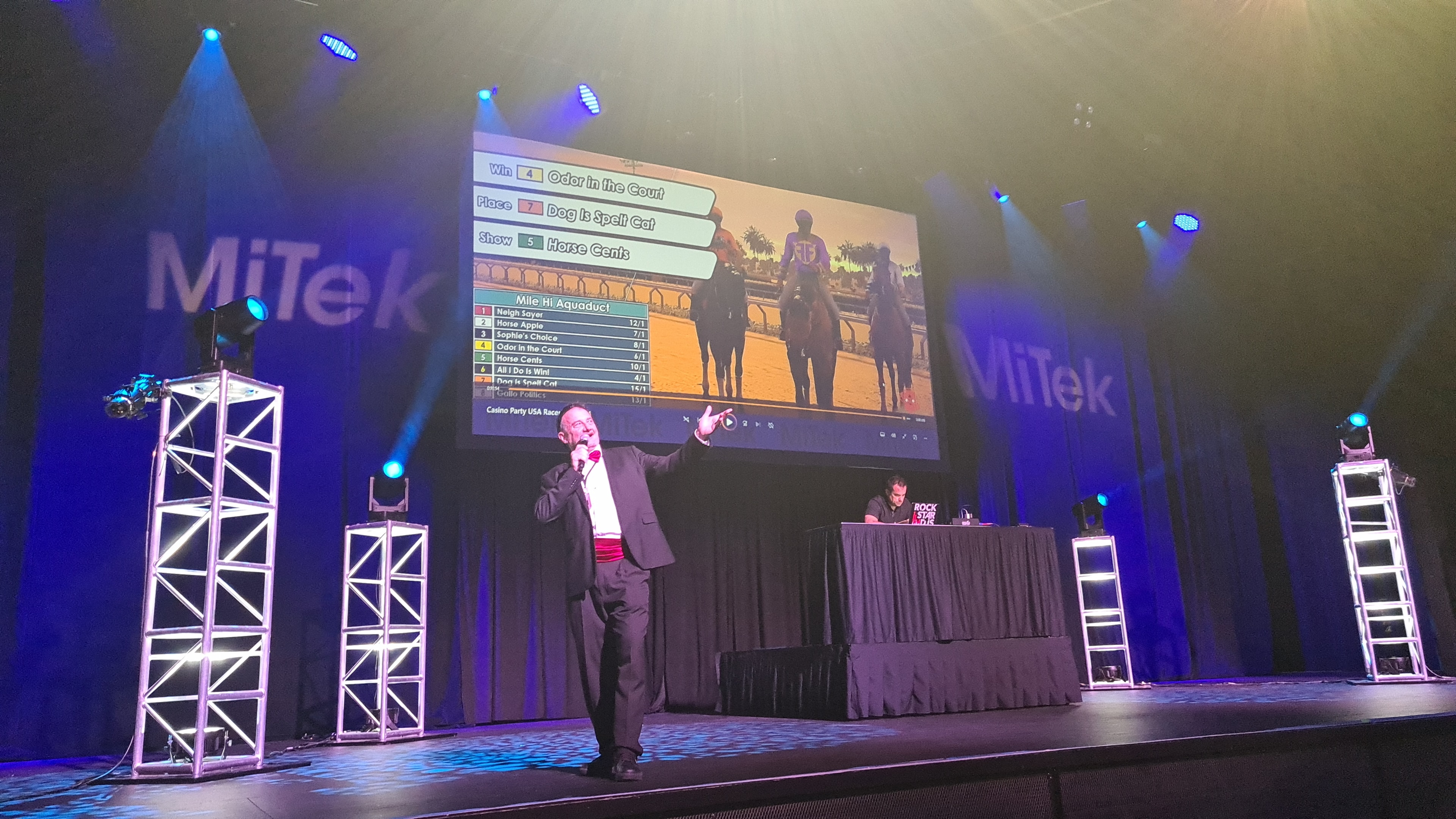 Casino Party USA race announcer on stage calling the virtual horse race at the MiTek corporate event with results displayed on the big screen